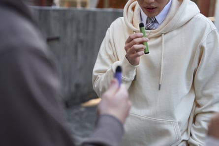 a boy in a cream hoodie smoking a vape in the playground
