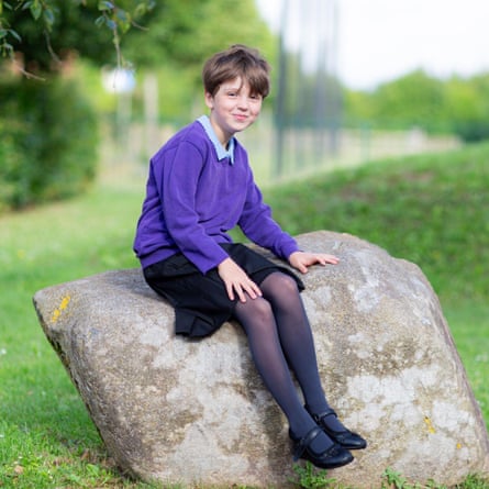 A young person sits on a rock with a school field and greenery in soft focus behind them. They are wearing a purple jumper with a light blue collar and navy skirt. They have medium denier navy tights and black one strap shoes on.