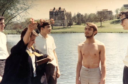 Group of young men at a lake with brick and limestone building on the far shore, with one man shirtless with his back to the water.