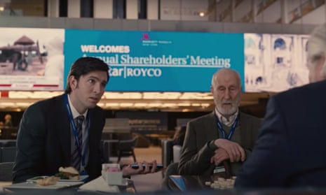 Video still of a meeting in the lobby of a conference centre or similar building. Greg has a look of consternation on his face as he is told something by a character just off screen, while Ewan, sitting next to him, has an unreadable expression