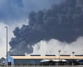 A thick cloud of black smoke rising about some port buildings