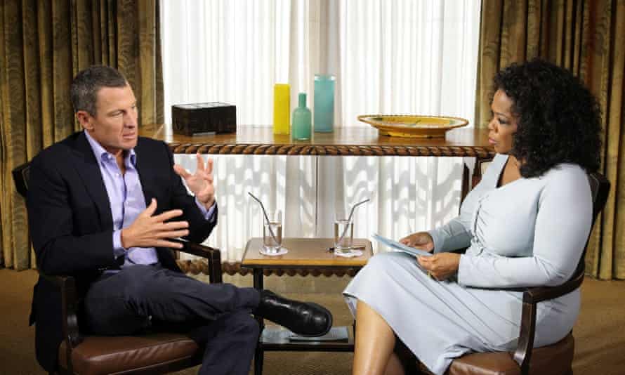 Armstrong in a dark suit and Winfrey in a light dress sit at a 45-degree angle to each other. They sit in a room with a sideboard at the back and two glasses of water on it.
