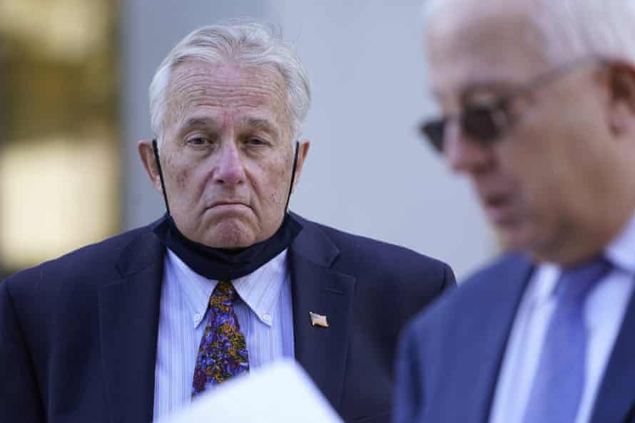 A man with his black mask pulled down around his chin listens as another man speaks in the foreground