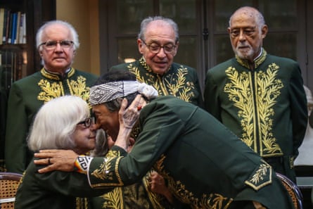 A woman and a man wearing a headband touch noses in front of three men. All are in ceremonial dress