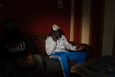 African man Yassin, wearing tracksuit, sits on the phone in a darkened living room.