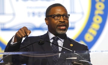 Middle-aged Black man with graying goatee and black-framed glasses in dark suit with lapel pin, speaks into microphone at clear lectern on stage, gesturing by pointing down with one finger.