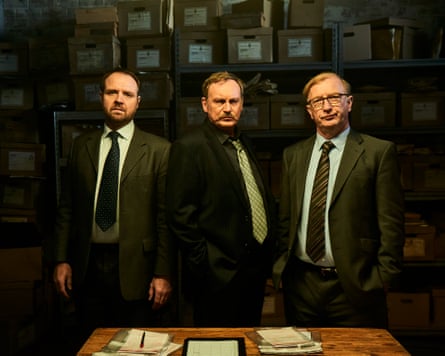 A moody publicity shot of Gareth John Bale, Philip Glenister and Steffan Rhodri standing in suits in a dim-lit evidence room