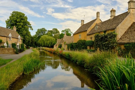 Yellow stone cottages alongside a river reflecting blue sky and clouds and lined by reeds