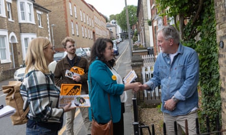 A Liberal Democrat candidate, Victoria Collins, canvasses with fellow party workers: they are in a residential street of brick houses talking to a man on his doorstep, holding leaflets and orange party signs. dqxikeidqkikdinv