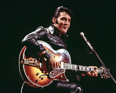 Elvis Presley, in a leather suit, plays an electric guitar on stage before a microphone.