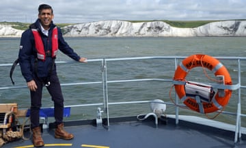 Rishi Sunnak on boat looking windblown. Life ring attached to railings that says 'HMC Seeker Border Force'. White cliffs in background
