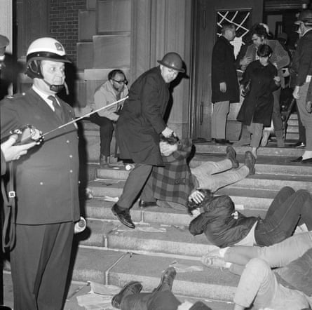 officer in helmet stands on steps while another man, standing, apparently pulls the hair of a person seated on the steps. others stand nearby or lie on the steps, in a black and white photo