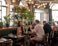 Diners sit at tables in an industrial-style restaurant with hanging Edison bulbs and potted plants