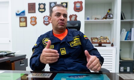 A Colombian man with buzzed brown hair, a bright orangeT-shirt collar showing under a dark blue uniform shirt with yellow stitching, sitting at a desk with many plaques on the white wall behind him.