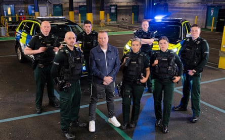 Nolan stands surrounded by seven police officers, in an underground car park with blue flashing lights