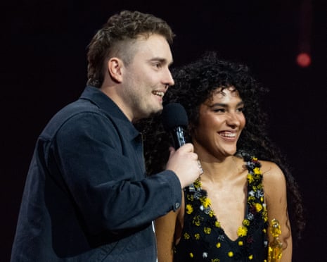 Sam Fender speaks into a microphone beside a smiling Olivia Dean on stage at the Brit Awards