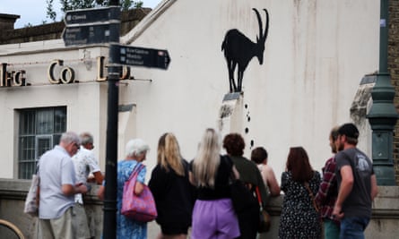 People stand behind a wall looking at a black spraypainted goat that appears to be balancing on a ledge with rocks falling below, on the side of a white building