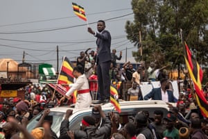 A crowd of people, some of them waving Ugandan flags, surround a car with a man standing on its roof who is also waving a small flag
