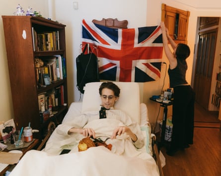 A man eats in a hospital bed at home, while a woman hangs a Union Jack flag behind him