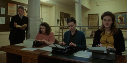 Three female typists work while a man stands reading.
