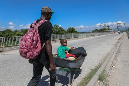 Man in cap and pink backpack pushes a 2- or 3-year old boy sitting on a few belongings in a black wheelbarrow on a long asphalt road.
