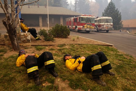 Three firefighters under smoky sky lay flat on lawn with hats covering faces or lean against a tree.