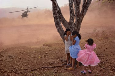 Three girls shield their eyes from dust beneath a tree as a military helicopter hovers overhead