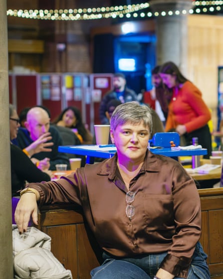 Elizabeth sits on a church pew; she has short grey hair, wears a brown satin shirt and blue jeans. Other attenders are in the background talking, reading and drinking coffee