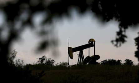 An oil pump jack silhouetted against the sky, in a picture framed by silhouetted tree branches and bushes