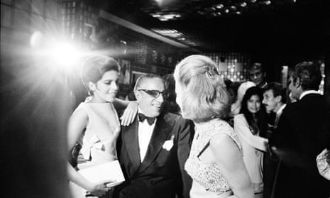 Aristotle Onassis in evening dress with one young woman leaning on his shoulder while he talks to another, with a bright light flaring in the top corner of the photo