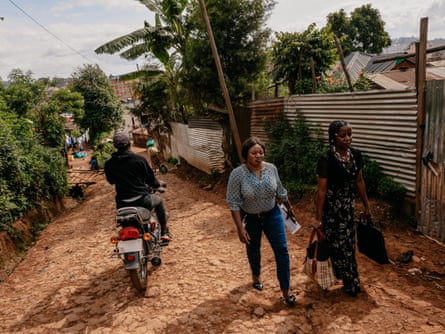 two women walk along a path of red soil and metal shacks