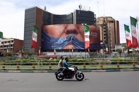 People on a motorbike ride near an anti-US billboard in Tehran with an image depicting US president Donald Trump.