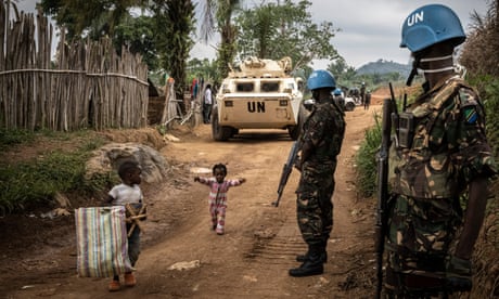 Tanzanian peacekeepers stand by a dirt road with two small children. A UN armoured personnel carrier stands in the background