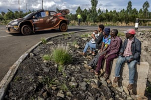 A family sit by the roadside, with a baby being fed as a rally car zooms past