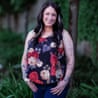 Shauna Castaner smiling at the camera, standing on a grass verge next to a path with her thumbs in her pockets, wearing a sleeveless floral top and blue jeans