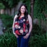 Shauna Castaner smiling at the camera, standing on a grass verge next to a path with her thumbs in her pockets, wearing a sleeveless floral top and blue jeans