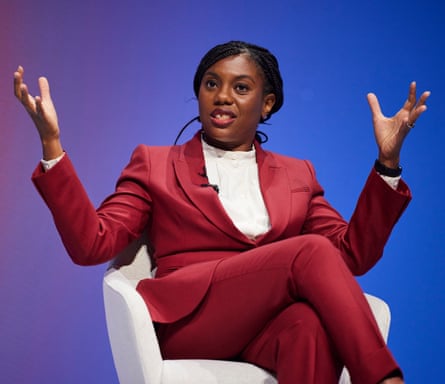 Kemi Badenoch, in a suit jacket and trousers, sits in a chair and holds up both hands as she speaks