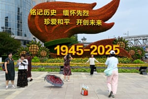 People take photographs in front of a flower installation that includes a large display shaped like a flaming torch and a sign that reads ‘1945-2025’
