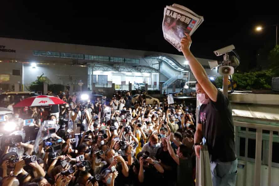 A man on a balcony holds up newspapers in front of a large crowd below, many of whom are press photographers