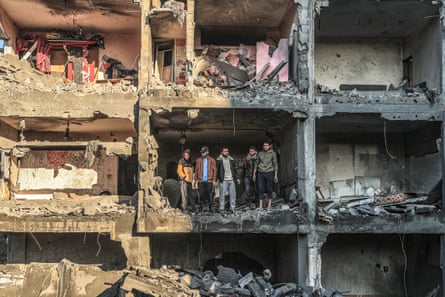 A photograph from the opposite side of the road of a group of men standing in a room of an apartment block that has entirely lost its exterior wall due to bomb damage.