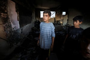 Two children, one with a bleeding gash to side of his temple, stand in darkened debris in room