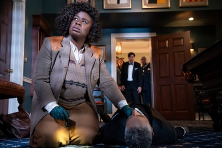 A black women in a tweed jacket and woolen tank top has her hand on teh neck of a white man who lies on the floor as if dead, a crowd watching on from a doorway