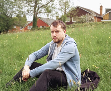 Leighton Allen poses for a picture sitting on a grassy slope with houses behind