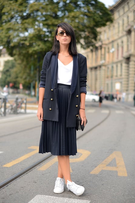 Model poses wearing a black skirt nd jacket, white Adidas trainers, top and and YSL sunglasses.