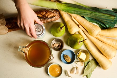 A hand arranges ingredients for parsnip and pear soup including pears, parsnips, a can of cider, garlic, leek, a cup of stock, bread, whole spices and butter