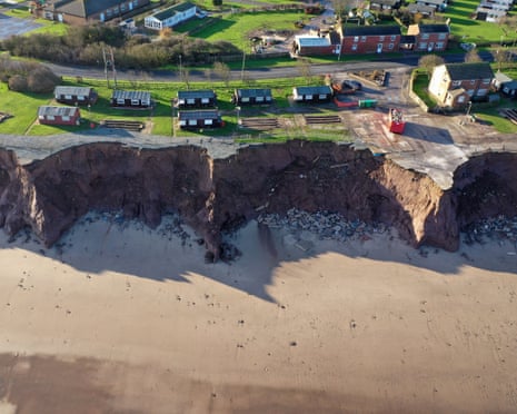 Aerial view of holiday chalets and houses near dramatic coastal erosion, with exposed rock face and beach below