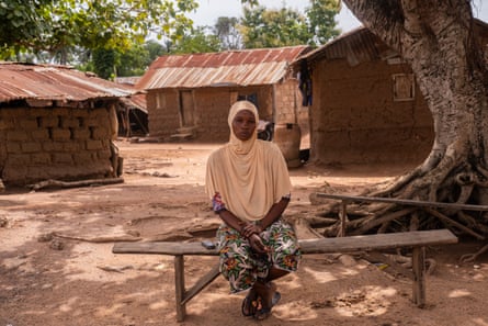 A woman wearing a headscarf sits on a bench under a tree in a village of mud-brick houses with corrugated roofs.