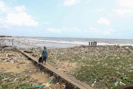 A man walks along a boardwalk on a deserted beach, with remnants of building structures rising from the sand.