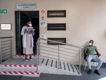 A woman in a white coat with a clipboard stands at the door of a clinic and a man sits on a chair outside. Both wear face masks