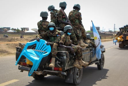 Men in military fatigues and helmets and carrying weapons are crowded on the back of a small truck. One sitting at the back holds two plastic chairs. The back of another such truck can be seen in front.